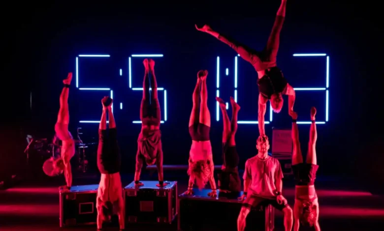 A imagem mostra uma apresentação de acrobacia em um palco iluminado com luzes em tons de vermelho e azul. Ao fundo, há um grande painel com números digitais luminosos que formam “55:113”. No centro do palco, oito acrobatas estão realizando paradas de mão, equilibrando-se sobre caixas pretas. À direita, um acrobata realiza um movimento avançado, apoiado apenas em uma das mãos na mão de outro acrobata sentado. O cenário é escuro, destacando os corpos iluminados dos artistas e o painel de números. À esquerda da imagem, há uma bateria, indicando a presença de música ao vivo na apresentação. A cena transmite força, equilíbrio e sincronização dos artistas.