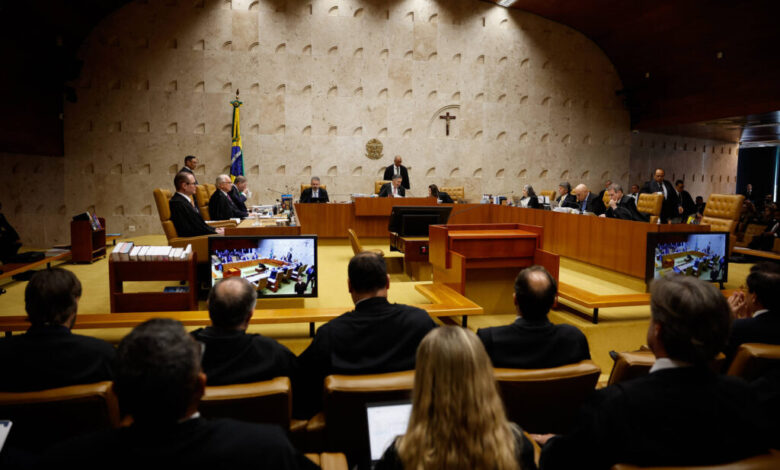 A imagem mostra uma sessão do Supremo Tribunal Federal do Brasil. No centro, há uma mesa com vários juízes e um presidente da sessão, todos em trajes formais. Ao fundo, há uma bandeira do Brasil e um crucifixo na parede. Na frente, há uma plateia com pessoas sentadas, algumas delas com trajes escuros. Em duas telas de projeção, é possível ver informações relacionadas à sessão.