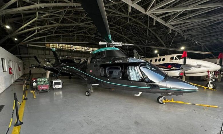 A imagem mostra um helicóptero preto com detalhes em verde, estacionado em um hangar. O helicóptero está em primeiro plano, com suas lâminas visíveis. Ao fundo, há um avião pequeno e outros equipamentos de aviação. O piso do hangar é de concreto e há algumas luzes acesas no teto.