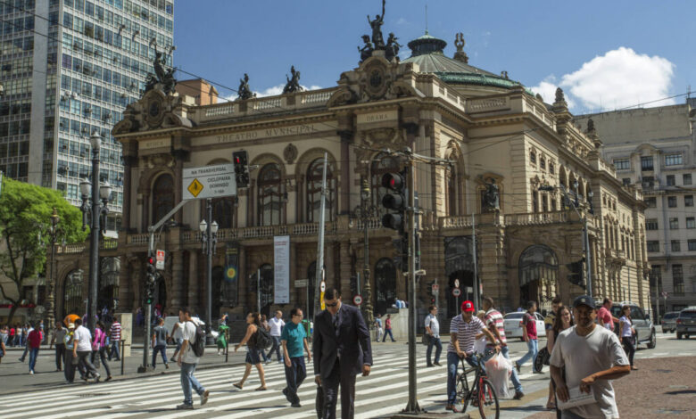 A imagem mostra o Teatro Municipal de São Paulo, um edifício histórico com uma arquitetura elaborada, localizado em uma área urbana movimentada. Na frente do teatro, há várias pessoas atravessando a rua em um cruzamento, algumas delas estão caminhando, enquanto outras andam de bicicleta. O céu está claro e ensolarado, e há prédios modernos ao fundo, contrastando com a arquitetura clássica do teatro.