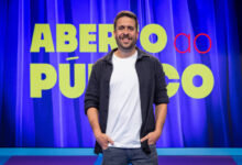 Um homem sorridente está em pé em frente a um fundo com cortinas azuis. Ele usa uma camiseta clara e uma jaqueta escura, com as mãos nos bolsos. Ao fundo, há um grande letreiro com as palavras