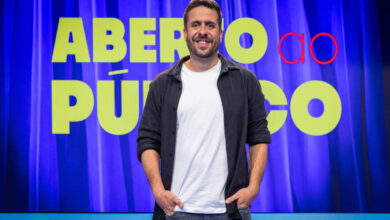 Um homem sorridente está em pé em frente a um fundo com cortinas azuis. Ele usa uma camiseta clara e uma jaqueta escura, com as mãos nos bolsos. Ao fundo, há um grande letreiro com as palavras