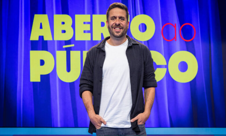 Um homem sorridente está em pé em frente a um fundo com cortinas azuis. Ele usa uma camiseta clara e uma jaqueta escura, com as mãos nos bolsos. Ao fundo, há um grande letreiro com as palavras