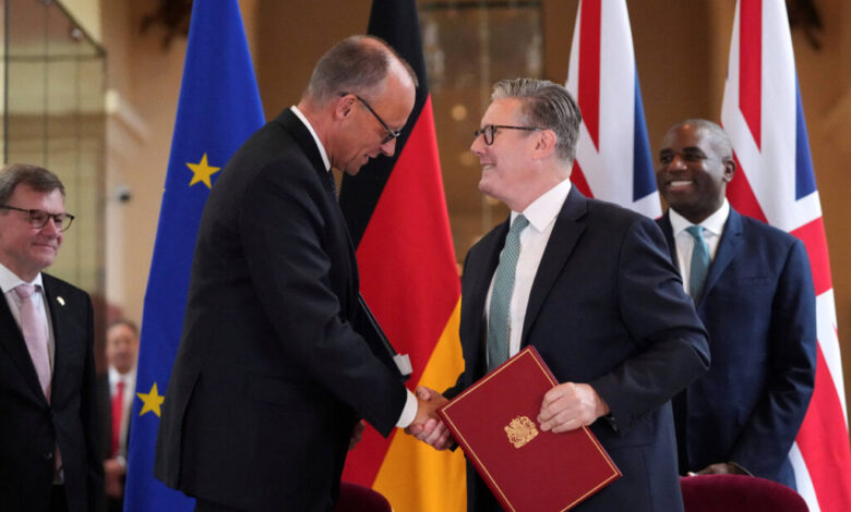 A imagem mostra uma cerimônia de assinatura de um acordo, onde duas pessoas estão se cumprimentando com um aperto de mão. Um deles está segurando um documento vermelho, enquanto o outro parece estar sorrindo. Ao fundo, há bandeiras da União Europeia, Alemanha, Reino Unido e outras. Há também outras pessoas presentes na cena, observando o evento.