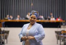 A imagem mostra uma mulher sorridente vestindo um blazer azul claro e um turbante colorido. Ela está em um ambiente formal, possivelmente uma sala de conferências ou um parlamento, com várias pessoas ao fundo sentadas em uma bancada. O fundo é composto por linhas verticais escuras e uma bancada de madeira.