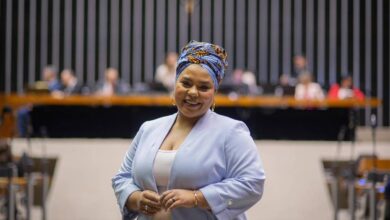 A imagem mostra uma mulher sorridente vestindo um blazer azul claro e um turbante colorido. Ela está em um ambiente formal, possivelmente uma sala de conferências ou um parlamento, com várias pessoas ao fundo sentadas em uma bancada. O fundo é composto por linhas verticais escuras e uma bancada de madeira.