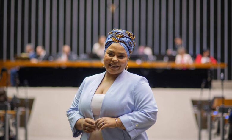 A imagem mostra uma mulher sorridente vestindo um blazer azul claro e um turbante colorido. Ela está em um ambiente formal, possivelmente uma sala de conferências ou um parlamento, com várias pessoas ao fundo sentadas em uma bancada. O fundo é composto por linhas verticais escuras e uma bancada de madeira.