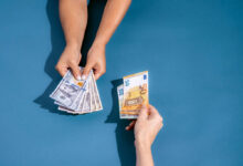 A imagem mostra duas mãos em um fundo azul. Uma mão está segurando várias notas de dólar, enquanto a outra mão está segurando uma nota de cinquenta euros. As mãos estão se aproximando, sugerindo uma troca de dinheiro.