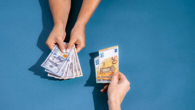 A imagem mostra duas mãos em um fundo azul. Uma mão está segurando várias notas de dólar, enquanto a outra mão está segurando uma nota de cinquenta euros. As mãos estão se aproximando, sugerindo uma troca de dinheiro.