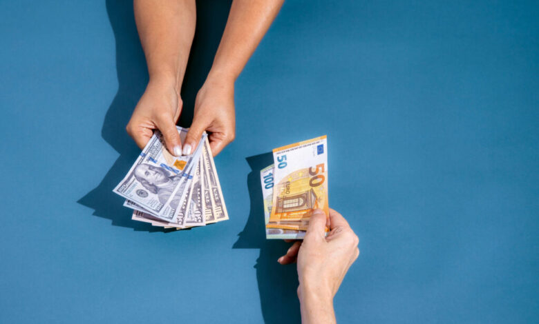 A imagem mostra duas mãos em um fundo azul. Uma mão está segurando várias notas de dólar, enquanto a outra mão está segurando uma nota de cinquenta euros. As mãos estão se aproximando, sugerindo uma troca de dinheiro.
