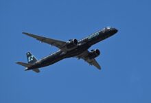 Um avião comercial está voando em um céu azul claro. O avião é de cor preta com detalhes em verde e possui a inscrição