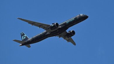 Um avião comercial está voando em um céu azul claro. O avião é de cor preta com detalhes em verde e possui a inscrição