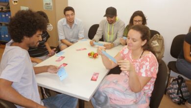 Seis pessoas sentadas em volta de uma mesa pegando papéis numa atividade educativa