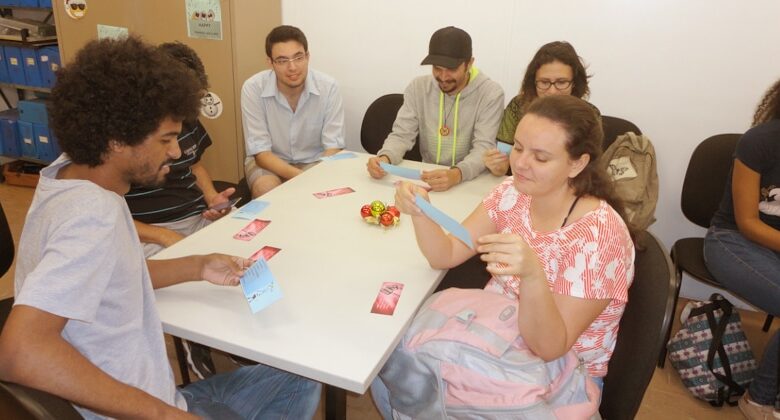 Seis pessoas sentadas em volta de uma mesa pegando papéis numa atividade educativa
