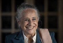 A imagem mostra uma mulher sorridente com cabelo grisalho preso em um coque. Ela está vestindo um paletó azul e uma camisa branca, com um lenço ou xale rosa sobre o ombro. O fundo é desfocado, destacando seu rosto iluminado e expressivo.