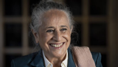 A imagem mostra uma mulher sorridente com cabelo grisalho preso em um coque. Ela está vestindo um paletó azul e uma camisa branca, com um lenço ou xale rosa sobre o ombro. O fundo é desfocado, destacando seu rosto iluminado e expressivo.