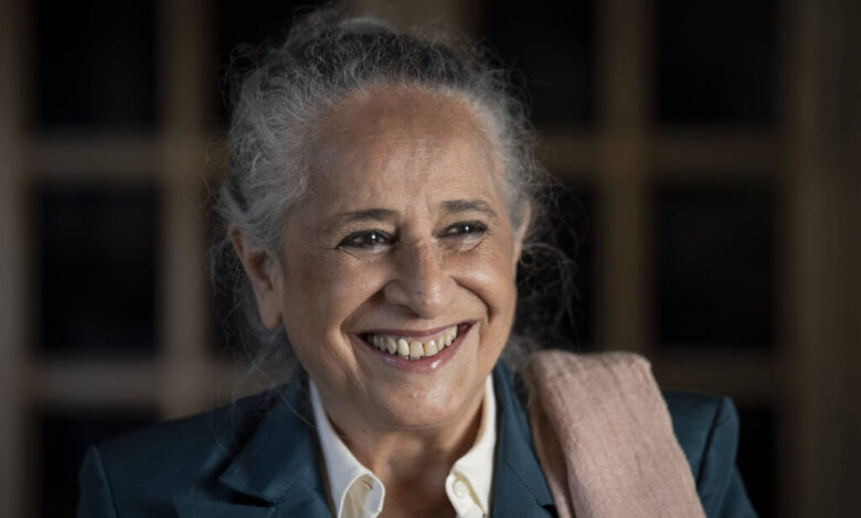 A imagem mostra uma mulher sorridente com cabelo grisalho preso em um coque. Ela está vestindo um paletó azul e uma camisa branca, com um lenço ou xale rosa sobre o ombro. O fundo é desfocado, destacando seu rosto iluminado e expressivo.