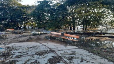 A imagem mostra uma área inundada, com água cobrindo parte do solo e detritos espalhados. Há bancos de madeira laranja visíveis, parcialmente submersos. Ao fundo, árvores estão presentes, e a luz do sol ilumina a cena, indicando que é durante o dia. A área parece ter sofrido os efeitos de uma enchente recente.