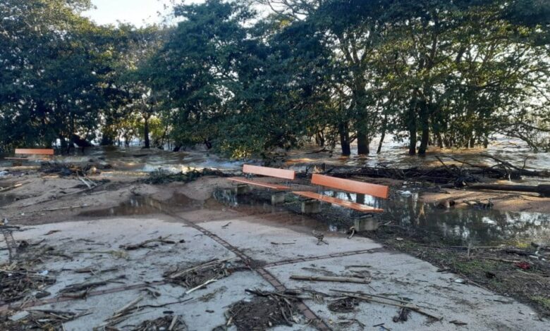A imagem mostra uma área inundada, com água cobrindo parte do solo e detritos espalhados. Há bancos de madeira laranja visíveis, parcialmente submersos. Ao fundo, árvores estão presentes, e a luz do sol ilumina a cena, indicando que é durante o dia. A área parece ter sofrido os efeitos de uma enchente recente.