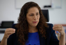 Uma mulher com cabelo cacheado e castanho claro está sentada em uma cadeira, gesticulando com as mãos enquanto fala. Ela usa uma blusa azul e um blazer escuro. O fundo é um ambiente de escritório, com uma mesa e um computador visíveis. A mulher parece estar em um momento de discussão ou entrevista.