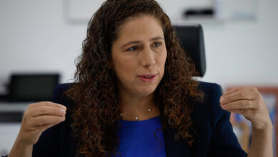Uma mulher com cabelo cacheado e castanho claro está sentada em uma cadeira, gesticulando com as mãos enquanto fala. Ela usa uma blusa azul e um blazer escuro. O fundo é um ambiente de escritório, com uma mesa e um computador visíveis. A mulher parece estar em um momento de discussão ou entrevista.