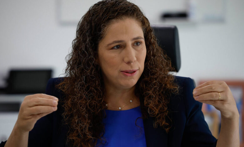 Uma mulher com cabelo cacheado e castanho claro está sentada em uma cadeira, gesticulando com as mãos enquanto fala. Ela usa uma blusa azul e um blazer escuro. O fundo é um ambiente de escritório, com uma mesa e um computador visíveis. A mulher parece estar em um momento de discussão ou entrevista.