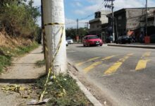 A imagem mostra um cruzamento de rua com um poste de luz à esquerda. O chão é de terra e asfalto, com algumas folhas secas ao redor. À direita, há um comércio com várias lojas visíveis e um carro vermelho passando pela rua. O céu está parcialmente nublado.