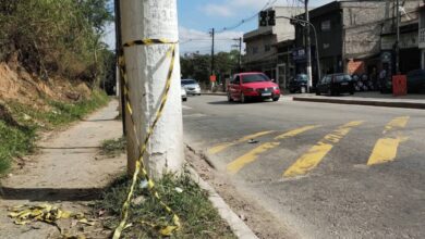 A imagem mostra um cruzamento de rua com um poste de luz à esquerda. O chão é de terra e asfalto, com algumas folhas secas ao redor. À direita, há um comércio com várias lojas visíveis e um carro vermelho passando pela rua. O céu está parcialmente nublado.