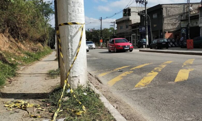 A imagem mostra um cruzamento de rua com um poste de luz à esquerda. O chão é de terra e asfalto, com algumas folhas secas ao redor. À direita, há um comércio com várias lojas visíveis e um carro vermelho passando pela rua. O céu está parcialmente nublado.