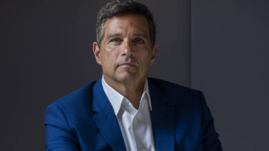 Um homem está sentado à mesa, vestindo um terno azul e uma camisa branca. Ele tem cabelo curto e grisalho, e está olhando para a câmera com uma expressão séria. A mesa reflete sua imagem, criando um efeito visual interessante. O fundo é de cor neutra, destacando o sujeito da imagem.