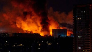 A imagem mostra um grande incêndio à noite, com chamas intensas e fumaça densa subindo em direção ao céu. No fundo, há edifícios iluminados pela luz do fogo, enquanto a parte inferior da imagem apresenta uma área urbana com várias luzes acesas. O céu está escuro, contrastando com o brilho laranja das chamas.