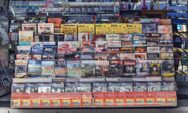 A imagem mostra uma banca de jornal com uma variedade de revistas e jornais expostos. A banca está organizada em prateleiras, com capas visíveis de diferentes publicações. À direita, uma mulher está de pé, usando uma máscara facial. O ambiente é iluminado e a banca é coberta por uma lona azul na parte superior.