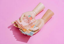 A imagem mostra um par de mãos de boneca segurando várias notas de dinheiro em cima de um fundo rosa. As notas são de diferentes valores e cores, destacando-se na composição.