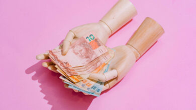 A imagem mostra um par de mãos de boneca segurando várias notas de dinheiro em cima de um fundo rosa. As notas são de diferentes valores e cores, destacando-se na composição.