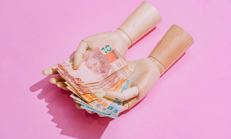 A imagem mostra um par de mãos de boneca segurando várias notas de dinheiro em cima de um fundo rosa. As notas são de diferentes valores e cores, destacando-se na composição.