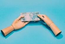 A imagem mostra duas mãos de madeira, uma de cada lado, segurando uma nota de 100 reais. O fundo é azul claro, destacando a nota que possui a figura de uma mulher e o valor impresso em destaque. As mãos estão posicionadas de forma a parecer que estão trocando a nota.