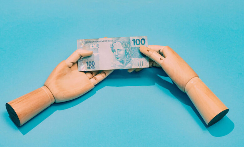 A imagem mostra duas mãos de madeira, uma de cada lado, segurando uma nota de 100 reais. O fundo é azul claro, destacando a nota que possui a figura de uma mulher e o valor impresso em destaque. As mãos estão posicionadas de forma a parecer que estão trocando a nota.