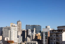 A imagem mostra uma vista panorâmica de um centro urbano, com diversos edifícios de diferentes tamanhos e estilos arquitetônicos. O céu está limpo e azul, e os prédios variam entre estruturas modernas e mais antigas, refletindo a diversidade arquitetônica da cidade.