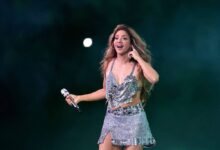 Uma artista está se apresentando ao vivo em um palco. Ela usa um vestido brilhante e curto, com detalhes que refletem a luz. A artista segura um microfone e parece estar cantando, com uma expressão animada e cabelo solto. O fundo é de um tom escuro, com uma luz difusa que destaca a figura da artista.
