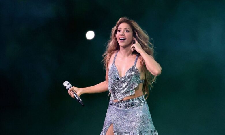 Uma artista está se apresentando ao vivo em um palco. Ela usa um vestido brilhante e curto, com detalhes que refletem a luz. A artista segura um microfone e parece estar cantando, com uma expressão animada e cabelo solto. O fundo é de um tom escuro, com uma luz difusa que destaca a figura da artista.