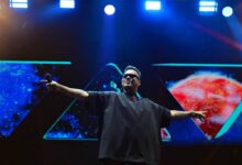 Um artista se apresenta em um palco iluminado, usando óculos escuros e uma camiseta larga. Ele está com os braços abertos, em uma pose de celebração ou interação com o público. Ao fundo, há um telão com imagens de espaço e planetas, criando um ambiente vibrante e dinâmico.