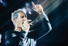 Um cantor está se apresentando em um palco, segurando um microfone e cantando. Ele está vestido com uma camisa de manga longa preta e tem cabelo grisalho. Raios de luz estão iluminando a cena, criando um efeito dramático ao fundo.