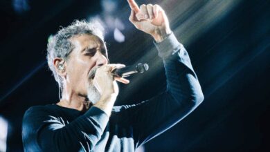 Um cantor está se apresentando em um palco, segurando um microfone e cantando. Ele está vestido com uma camisa de manga longa preta e tem cabelo grisalho. Raios de luz estão iluminando a cena, criando um efeito dramático ao fundo.