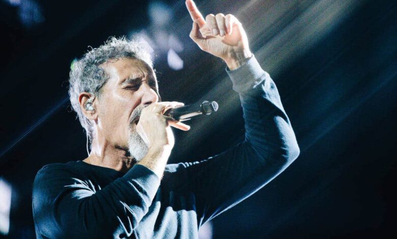 Um cantor está se apresentando em um palco, segurando um microfone e cantando. Ele está vestido com uma camisa de manga longa preta e tem cabelo grisalho. Raios de luz estão iluminando a cena, criando um efeito dramático ao fundo.