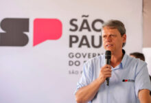 Um homem está falando ao microfone em um evento. Ele usa uma camisa azul clara e está em frente a um fundo com a logo do governo de São Paulo, que inclui as letras