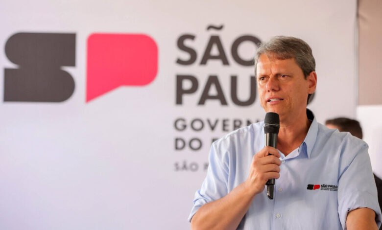Um homem está falando ao microfone em um evento. Ele usa uma camisa azul clara e está em frente a um fundo com a logo do governo de São Paulo, que inclui as letras