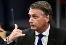 Um homem com cabelo castanho e liso, usando um terno preto e uma gravata azul, está em uma reunião. Ele está olhando para o lado e fazendo um gesto com a mão, como se estivesse fazendo um sinal de telefone. Ao fundo, há uma mesa e outros elementos de um ambiente de reunião.