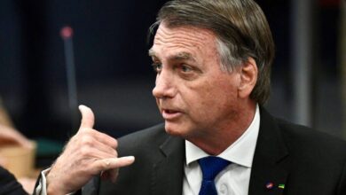 Um homem com cabelo castanho e liso, usando um terno preto e uma gravata azul, está em uma reunião. Ele está olhando para o lado e fazendo um gesto com a mão, como se estivesse fazendo um sinal de telefone. Ao fundo, há uma mesa e outros elementos de um ambiente de reunião.
