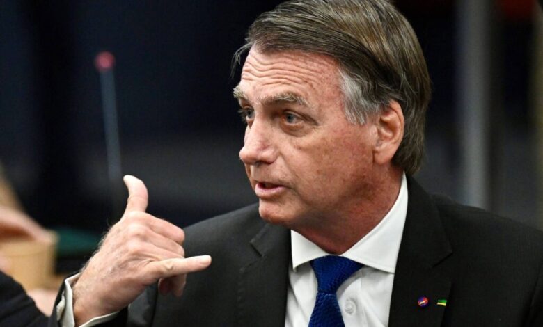 Um homem com cabelo castanho e liso, usando um terno preto e uma gravata azul, está em uma reunião. Ele está olhando para o lado e fazendo um gesto com a mão, como se estivesse fazendo um sinal de telefone. Ao fundo, há uma mesa e outros elementos de um ambiente de reunião.