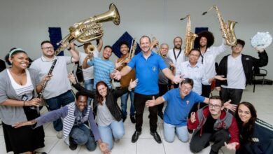 A imagem mostra um grupo grande e animado de músicos, muitos segurando seus instrumentos, sorrindo para a câmera. Há uma mistura de instrumentos de sopro, incluindo trompetes, saxofones, e um tuba. Um homem de camisa azul está no centro da foto, com os braços abertos, como se estivesse conduzindo ou liderando o grupo. As pessoas ao redor dele parecem ser de diversas idades e etnias. A atmosfera é descontraída e festiva, sugerindo uma reunião de banda ou um ensaio. O fundo é simples, uma parede branca com um pouco de decoração, indicando um ambiente interno como um salão de ensaio ou uma sala de aula. A iluminação é uniforme, sem sombras fortes, contribuindo para a clareza da imagem.